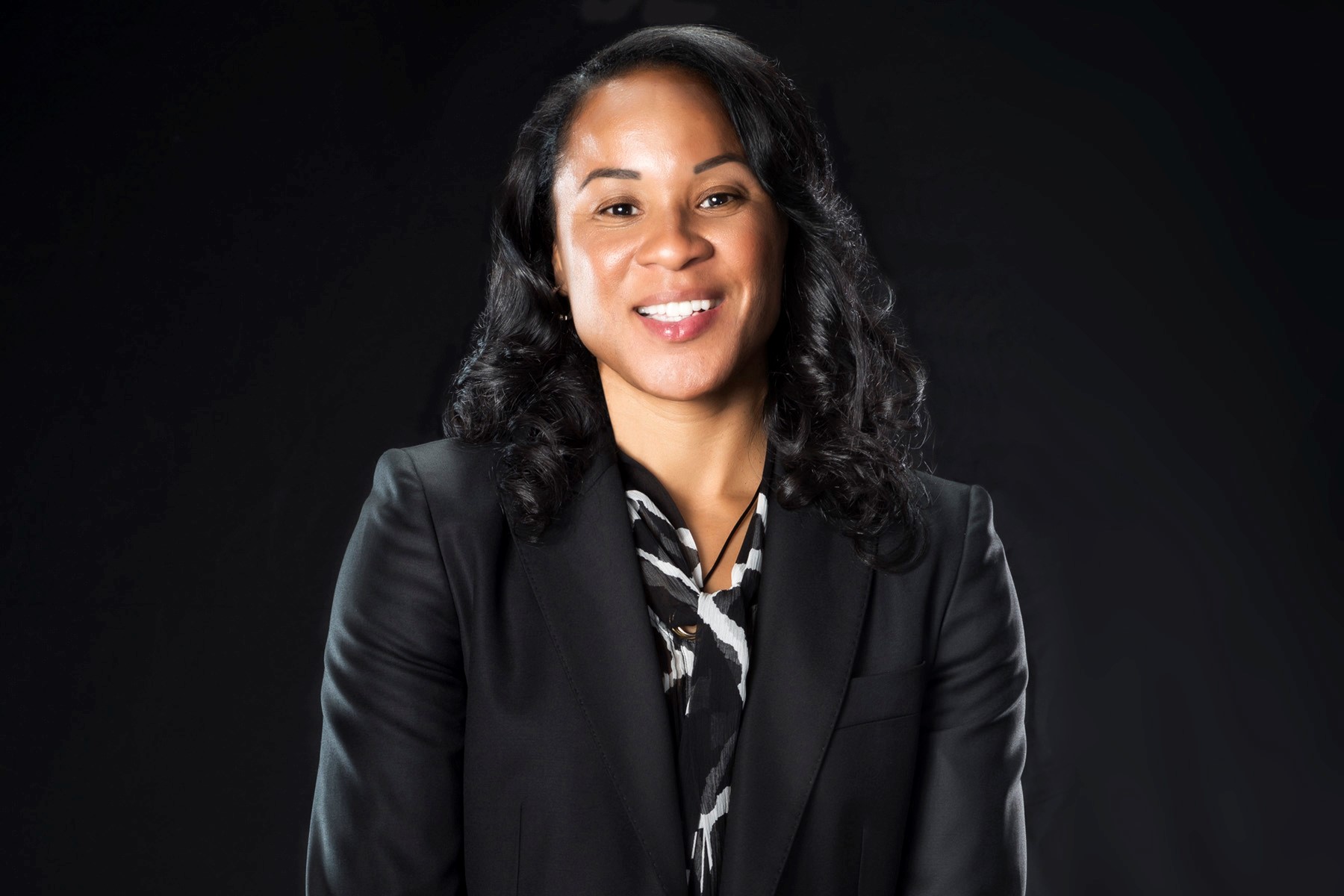 A photo of USC Women's Basketball Coach Dawn Staley. Coach Staley is photographed against a black background in a black blazer and black and white blouse.