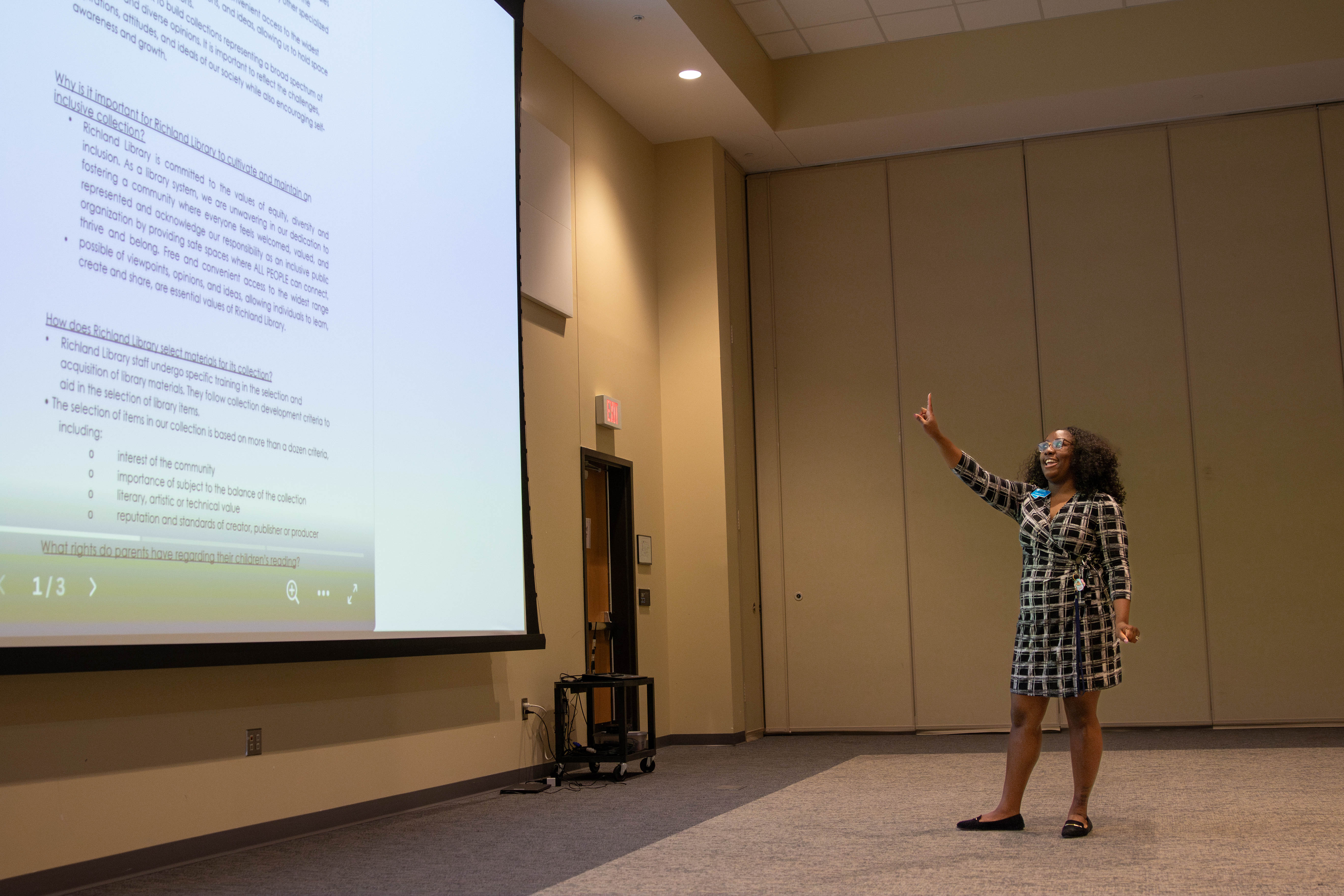Black woman in dress pointing to a presentation screen in a high ceiling conference room